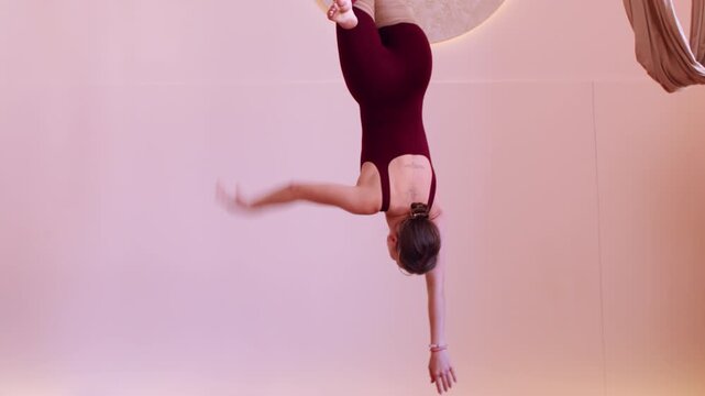 Aerial yoga. Body stability. Woman holding on wrapped fabric hammock training muscle strength in balance rotation pose in gym interior.