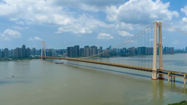 Aerial timelapse of Yangsigang Yangtze River Bridge in Wuhan, Hubei China, sunny day with blue sky and white clouds, city skyline along Yangtze river, golden double-deck suspension bridge, urban river