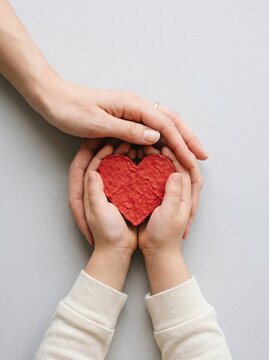 Red paper heart held in hands of mother and child, family love and care concept, charity and support symbol on gray background