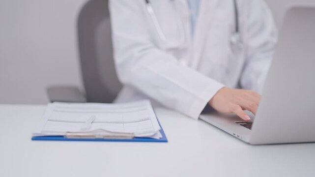 Professional doctor wearing lab coat and stethoscope using laptop computer and taking report notes on notebook at desk in hospital clinic office, medical technology and healthcare concept