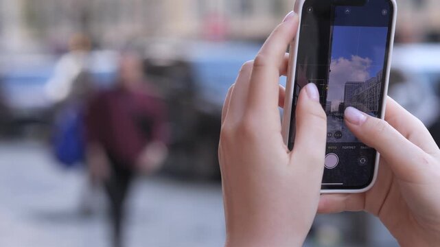 Hand holds smartphone, capturing street scene. Focus on phone screen, blurred background. Urban life captured through camera lens. Ideal for social media, travel, or photography content