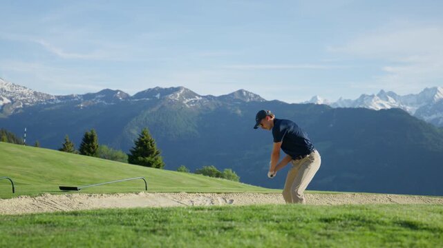 Skilled golfer blasts a shot from a sand bunker on a mountain golf course in Verbier, Switzerland. Slow motion sand spray with dramatic alpine peaks in the background.