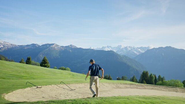 Wide cinematic shot of a golfer walking toward a sand bunker on a mountain golf course in Verbier, Switzerland. Expansive alpine scenery and dramatic Swiss peaks create a premium outdoor sports settin