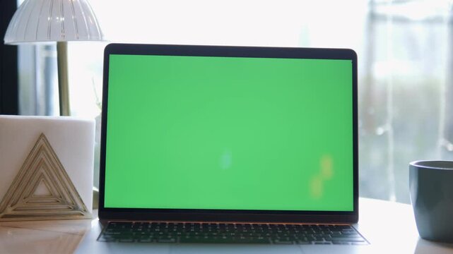 Laptop with green screen sits on wooden desk. Soft light comes from window behind. Modern lamp and mug beside laptop. Minimalist workspace with clean lines and calm vibe. Perfect for video editing