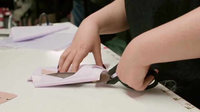 Dressmaker service. Clothing production. Female seamstress hand cutting white fabric with scissors figure paper pattern draft on studio desk.