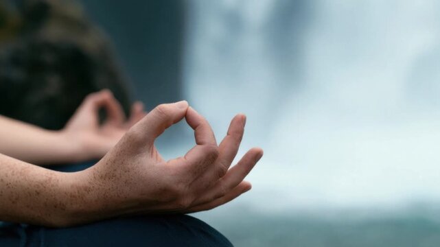 Close-up of relaxed hand forming meditation gesture with index finger and thumb touching during peaceful outdoor mindfulness practice near waterfall