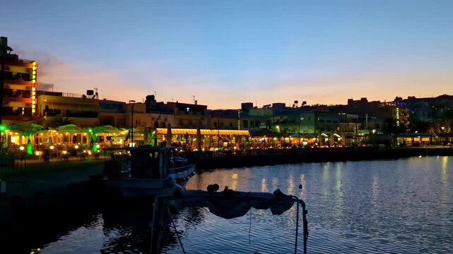 Colorful harborfront cafes and apartments glow at blue hour as small boats sit moored on the calm Sitia waterfront.