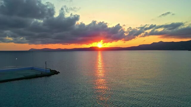 Fiery sunset light reflects across quiet coastal water as a long concrete pier extends into the sea under low clouds.