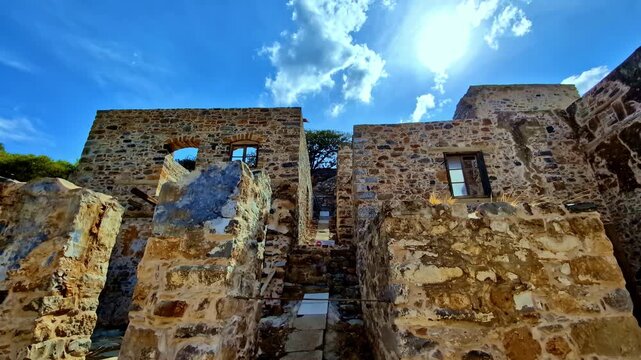 Weathered stone walls of Spinalonga fortress rise under a bright midday sun as the camera looks up through the ruined courtyard.