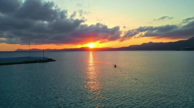 Lone kayaker paddles across quiet open water toward a vivid sunset as clouds drift above distant mountains.