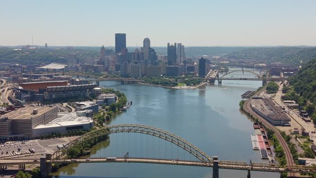 Aerial drone perspective tracking along a wide river toward a major highway bridge and the urban downtown city skyline on a clear sunny day