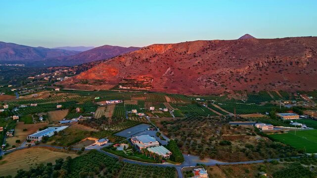 Sunset light colors the Heraklion hillside while a drone passes above orchards, fields and scattered farmhouses below.