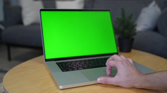 a person's hand using a silver laptop touchpad with a blank green screen. The computer sits on a round wooden table in a bright modern living room with a blurred sofa background.