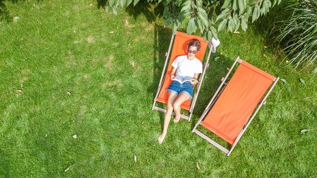 Young girl relaxes and reading book in summer garden in sunbed deckchair on grass, relaxing outdoors in green park on weekend, aerial drone view from above
