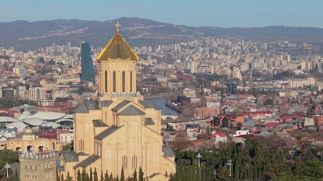 Drone orbit around the Holy Trinity Cathedral of Tbilisi, featuring the golden dome and panoramic cityscape in the background.