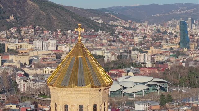 Drone shot of the Holy Trinity Cathedral dome in Tbilisi revealing the Kura River, riverbank buildings, and a nearby bridge under daylight.