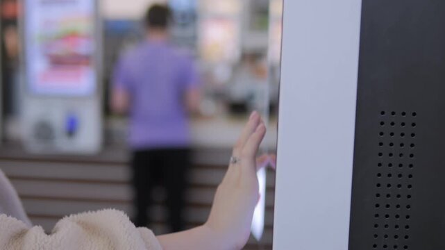 Close-up of hand interacting with modern touchscreen kiosk. Background shows blurred retail space and another person walking. Focus on user's gesture and kiosk interface. Suitable for retail, tech