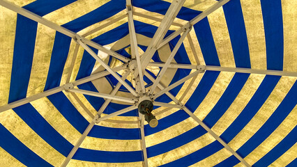 Close up of Symmetrical bottom view of a blue and yellow striped beach umbrella showcasing its radial geometric pattern against the sky. This colorful outdoor parasol creates an eye-catching abstract  © RahmawatiNur