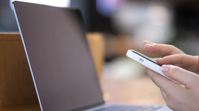 Person holds smartphone while laptop rests nearby. Focus on hands and device interaction. Suggests multitasking, digital communication, or remote work