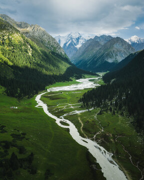 Aerial view of Boris Yeltsin Peak towering over a lush green valley with a winding river and dense coniferous forests in Jety-Oguz, Issyk-Kul Region, Kyrgyzstan.