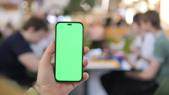 Hand holds smartphone with green screen. Background shows blurred cafe patrons. People appear relaxed and focused on their own tasks. Ideal for app demos, social media templates, or mockups