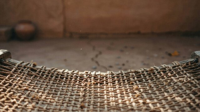 Cott bed made of woven ropes stands on a dirt floor inside a traditional mud house with a clay pot in background.