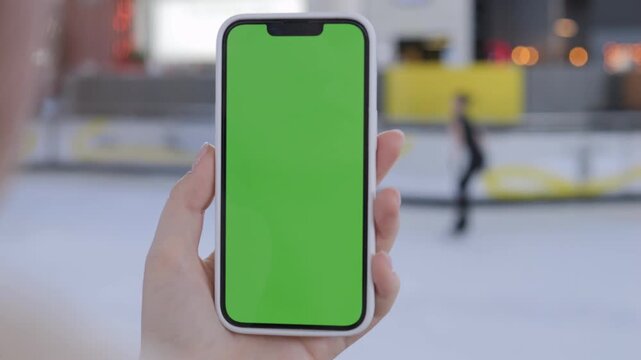 Hand holds smartphone with green screen. Ice rink in background. Person skates nearby. Useful for app demos or social media content. Focus on device, not surroundings