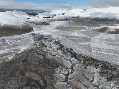 Aerial view of the snow-covered Piano Grande near Castelluccio, featuring winding streams and dramatic mountain peaks in the background in Castelluccio, Umbria, Italy.