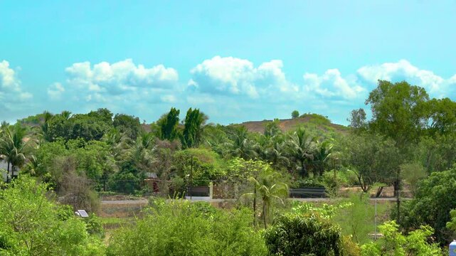 Slow zoom out revealing a wider tropical Indian village scene with dense greenery, palm-covered hills, roadside traffic moving in both directions, and open sky.