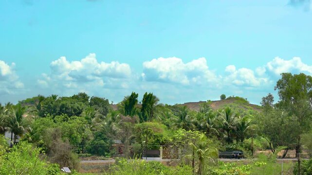 Static shot of a lush tropical Indian village landscape with palm trees, green hills, a roadside scene, and moving traffic including cars, bikes, trucks, and other vehicles under a bright blue sky wit