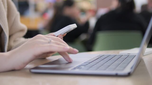 Close-up of hands using both laptop and smartphone. Person multitasks while seated at a wooden table. Blurred background suggests cafe or public workspace. Focus on fingers typing and swiping