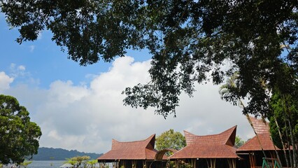 Traditional wooden pavilions beneath a lush tree canopy with blue sky and clouds, creating a peaceful tropical landscape perfect for travel, architecture, and nature themes. © mrbudiarto
