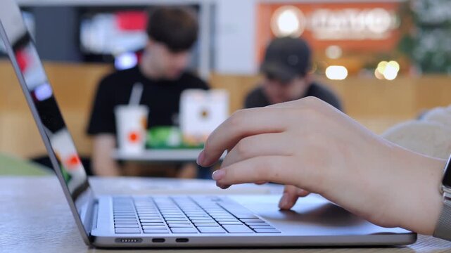 Close-up of hand typing on laptop keyboard. Focused fingers show concentration and productivity. Background shows blurred people in casual setting. Ideal for work, study