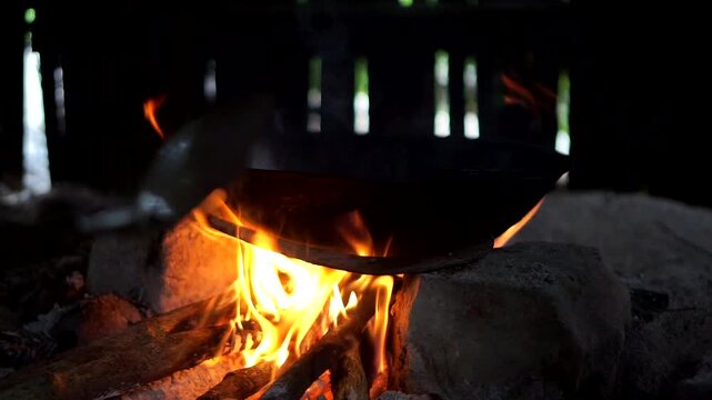 A traditional black wok placed on a simple stone hearth with blazing fire from burning firewood, showcasing an ancient cooking method inside a rural hut.