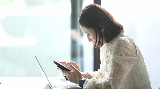 Soft light filters through large windows. Woman sits, focused on her tablet. Laptop rests beside her, open. She taps gently, absorbed in work. Dressed in light, textured blouse