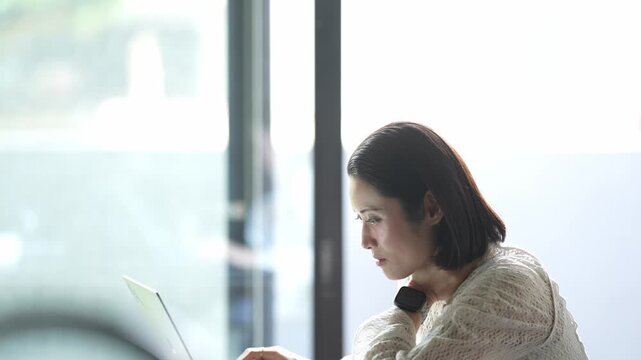 Soft light fills a quiet workspace. Woman works calmly, focused on her laptop. Warm sweater adds comfort to her seated posture. Natural daylight enhances her serene concentration