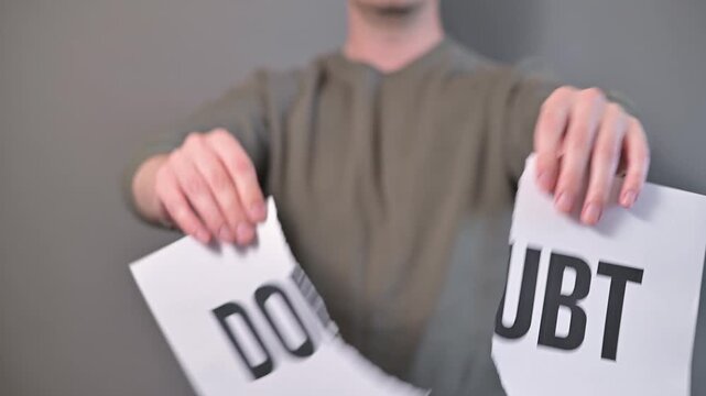 A person rips a sign that reads Doubt in half, showing determination and strength. This action signifies overcoming obstacles and embracing self-belief in a neutral setting