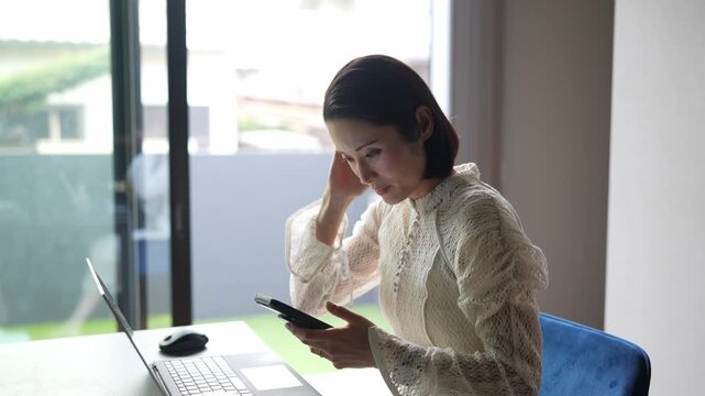 Woman focused on laptop, holding phone. Soft natural light illuminates her workspace. She rests head on hand, appearing tired or stressed. Ideal for remote work, digital fatigue, or daily grind themes