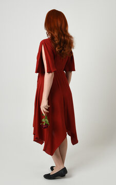Full length portrait of beautiful redhead model wearing elegant red modern dress. standing pose, walking away facing backwards, holding a romantic red rose, isolated figure studio background
