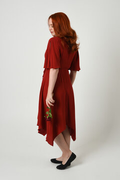 Full length portrait of beautiful redhead model wearing elegant red modern dress. standing pose, walking away facing backwards, holding a romantic red rose, isolated figure studio background