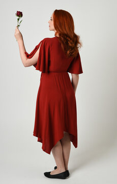 Full length portrait of beautiful redhead model wearing elegant red modern dress. standing pose, walking away facing backwards, holding a romantic red rose, isolated figure studio background