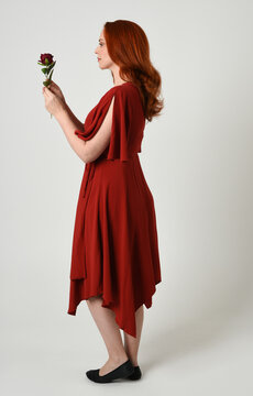 Full length portrait of beautiful redhead model wearing elegant red modern dress. standing pose, walking away facing backwards, holding a romantic red rose, isolated figure studio background