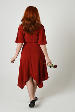 Full length portrait of beautiful redhead model wearing elegant red modern dress. standing pose, walking away facing backwards, holding a romantic red rose, isolated figure studio background