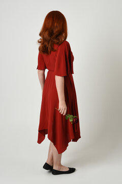 Full length portrait of beautiful redhead model wearing elegant red modern dress. standing pose, walking away facing backwards, holding a romantic red rose, isolated figure studio background