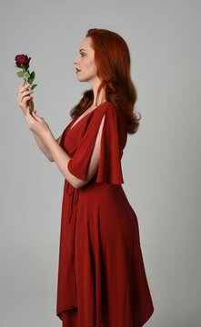close up portrait of beautiful model with long red hair, wearing an elegant red modern dress. standing pose holding a romantic red rose, isolated figure on studio background.
