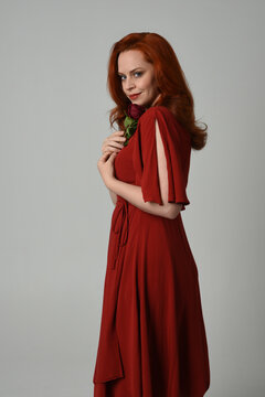 close up portrait of beautiful model with long red hair, wearing an elegant red modern dress. standing pose holding a romantic red rose, isolated figure on studio background.
