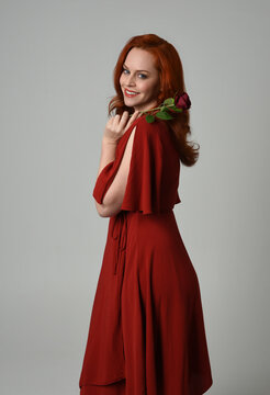 close up portrait of beautiful model with long red hair, wearing an elegant red modern dress. standing pose holding a romantic red rose, isolated figure on studio background.
