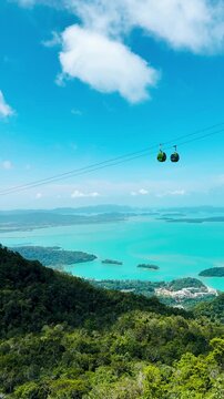 4K Vertical shot of cable car above the trees of forest and in front of turquoise blue colored sea and sky with clouds at Langkawi, Malaysia. Scenic travel background. Summer vacation concept.