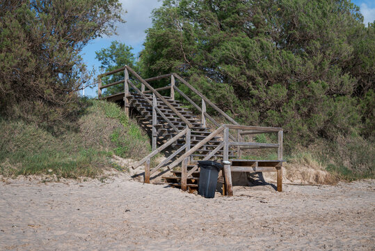 Wooden access stairs leading to a sandy beach in Costa Ballena, Chipiona, C&aacute;diz, Spain, surrounded by coastal vegetation under a partly cloudy sky, representing seaside access and summer tourism.