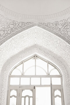Ornate white plaster carving surrounding a pointed arch window inside Muhammad Amin Khan Madrasah in Khiva. Intricate Islamic arabesques and floral patterns create an ethereal high-key composition.
